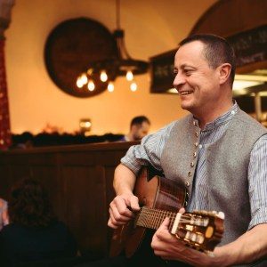 Man playing guitar and smiling in a cozy restaurant setting with warm lighting.
