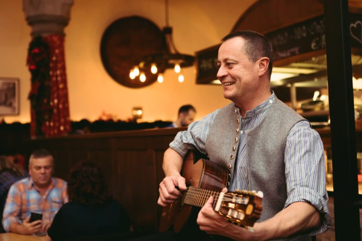 Man playing guitar and smiling in a cozy restaurant setting with warm lighting.