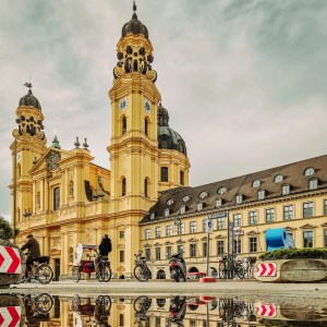 Historic yellow building with clock towers, bikes, and sky reflection in puddle.