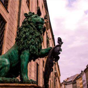 Green lion statue with shield in front of historic buildings under a cloudy sky.