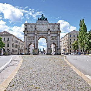 Triumphal arch on a wide street, flanked by trees and historic buildings under a blue sky.