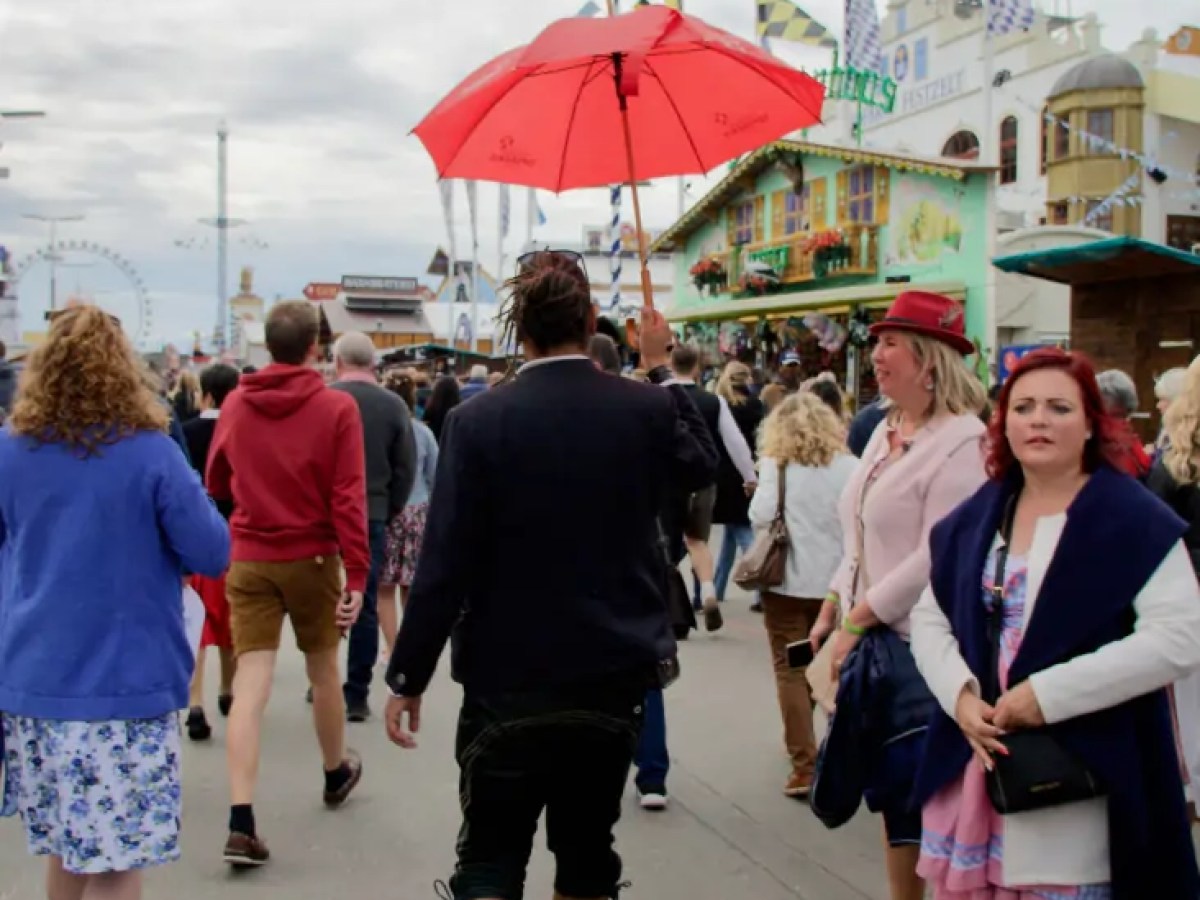 Crowd walking at a fairground; person with a red umbrella, amusement rides and festive buildings in the background.