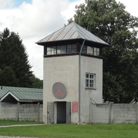 Gray concrete watchtower with a metal roof beside a green-roofed building, surrounded by trees and a fence.