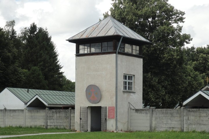 Gray concrete watchtower with a metal roof beside a green-roofed building, surrounded by trees and a fence.