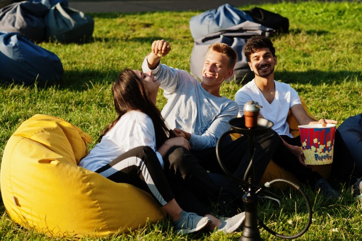 Three friends laughing on bean bags in a grassy area with popcorn and a hookah nearby.