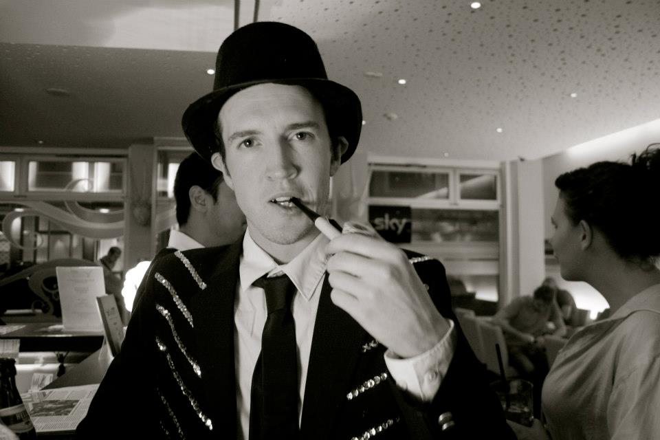 Man in a top hat and suit jacket with a pipe, sitting at a bar in a dimly lit room.