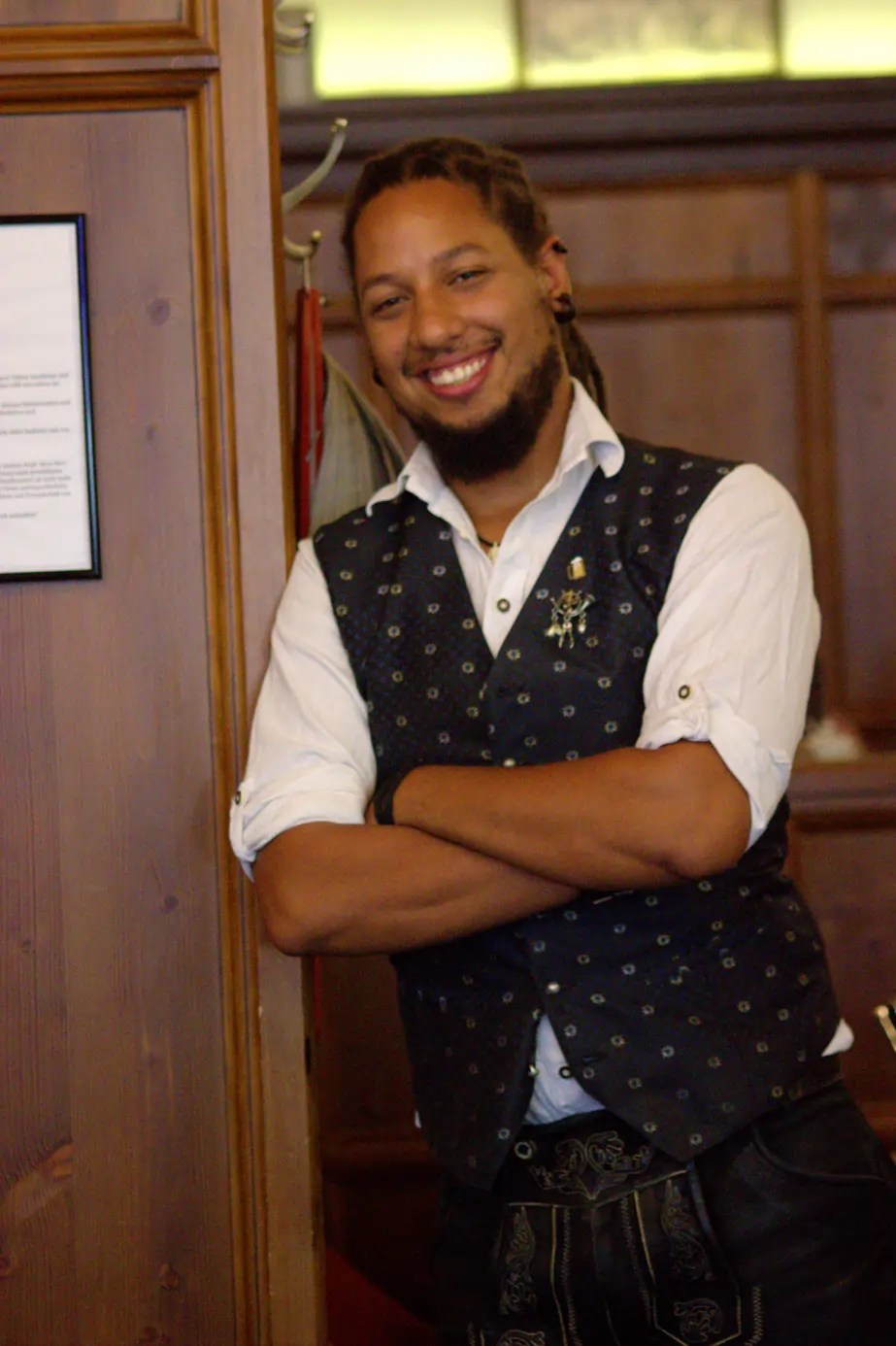 Person with a beard smiling, wearing a patterned vest and white shirt, standing in a wooden interior.