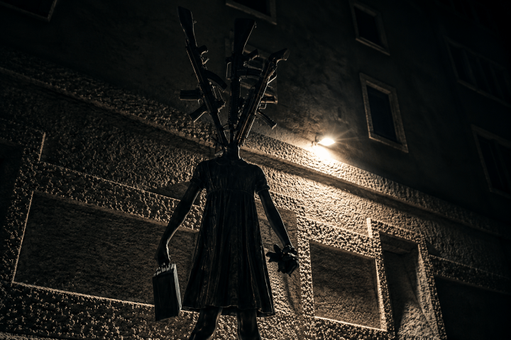 Dark statue of figure with branch-like head holding a bag, under dim streetlight.