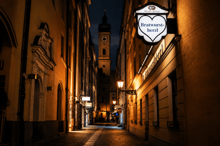 Narrow cobblestone street at night with lit signs and a distant church tower.
